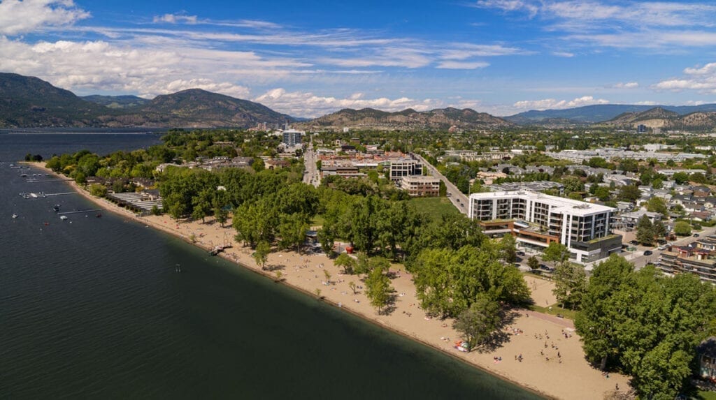 An aerial view of The Shore Kelowna accommodations on Okanagan Lake with Gyro beach in the foreground on a sunny day
