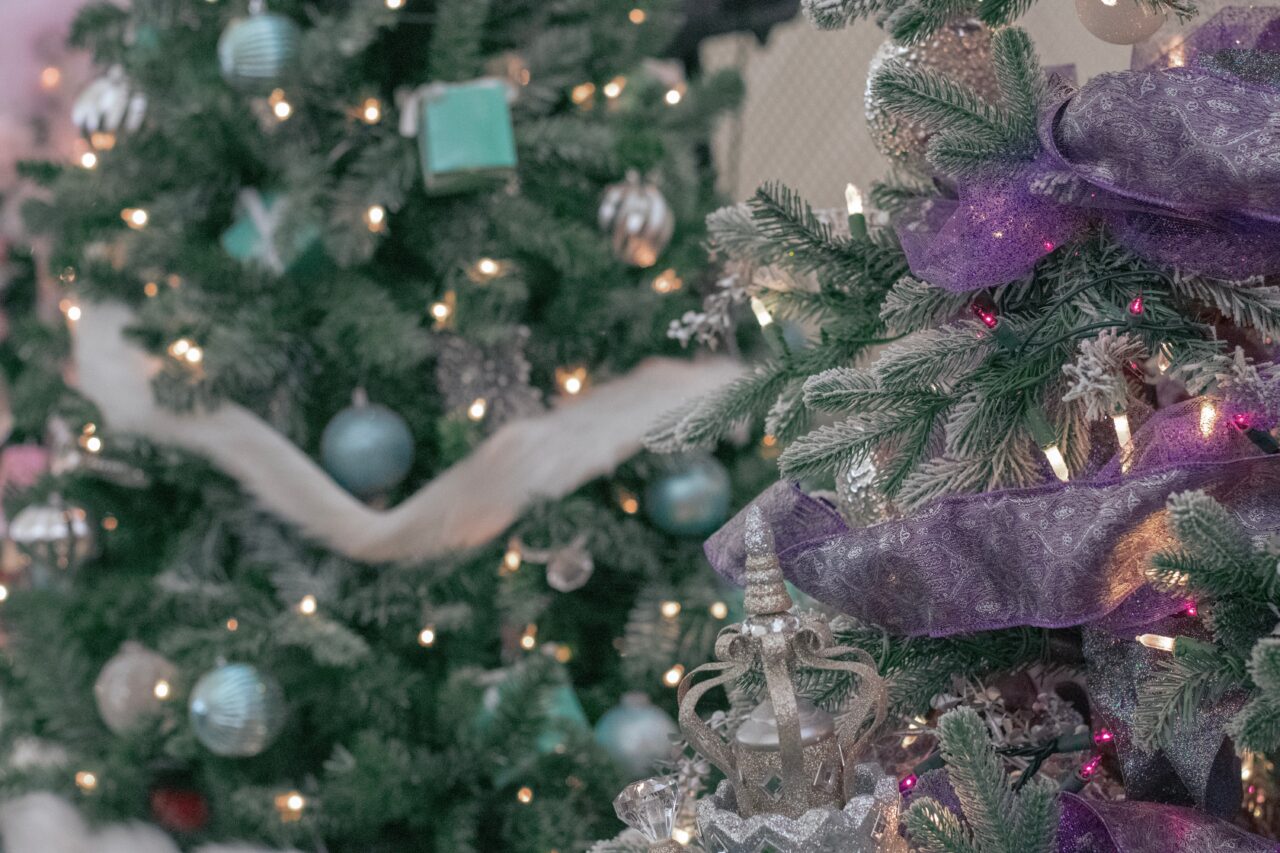 closeup of a christmas tree with purple ribbon and silver ornaments, and a christmas tree in the background with silver ribbon and teal ornaments