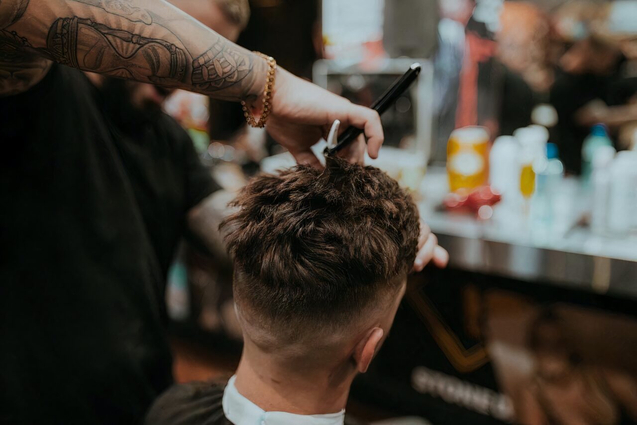 a man getting a haircut at a kelowna barber shop by a man with arm tattoo sleeve and a gold chain watch