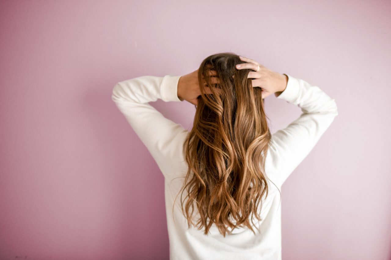 a woman facing a pink wall wearing a white sweater and holding her hair done at a kelowna hair salon