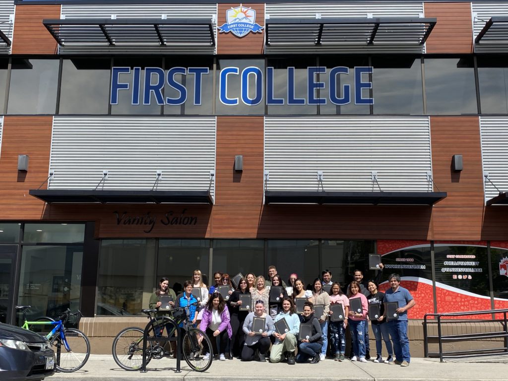 an exterior view of Kelowna First College, with graduates holding their diplomas, posing as a group at the entrance