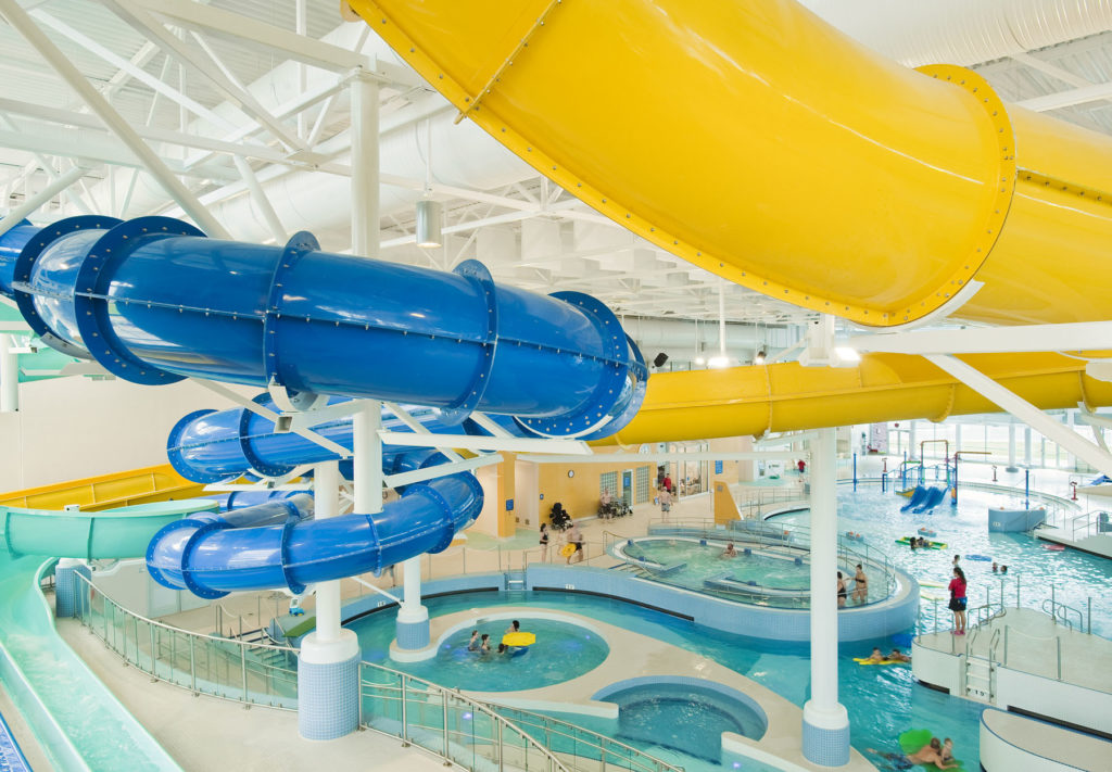 a view from the top of the waterslides at the Kelowna H2O aquatic centre, looking down on the various pools with families and staff members below.