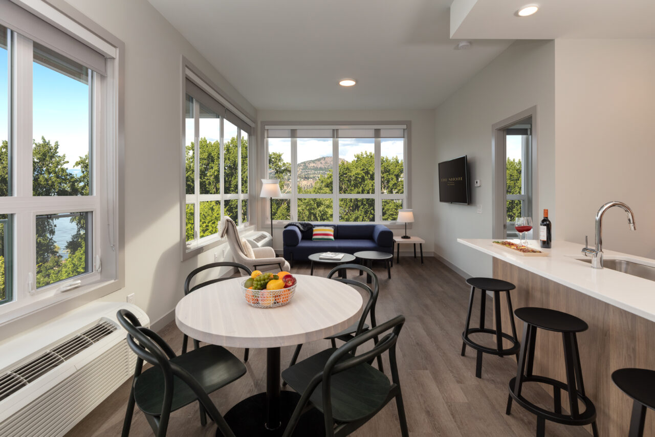 a suite interior at The Shore kelowna, showing a kitchen table, island bar with stools, and a living room area with a tv