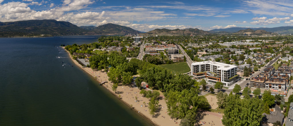 an exterior view of The Shore Kelowna showing the Pandosy neighbourhood and downtown kelowna in the background, with Gyro beach and Okanagan lake in the foreground