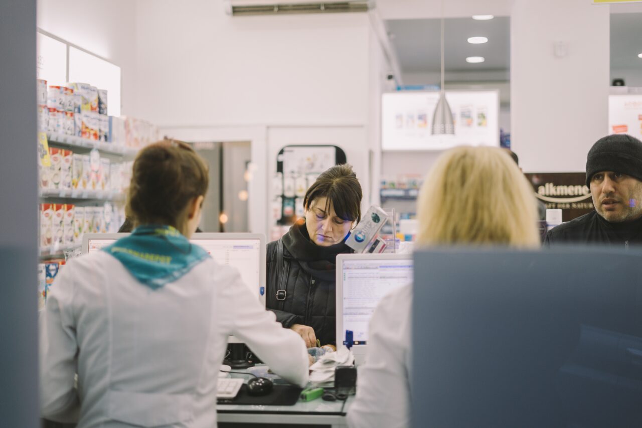 two customers stand behind a pharmacy counter to get prescriptions from two pharmacy assistants behind their computers in white lab coats