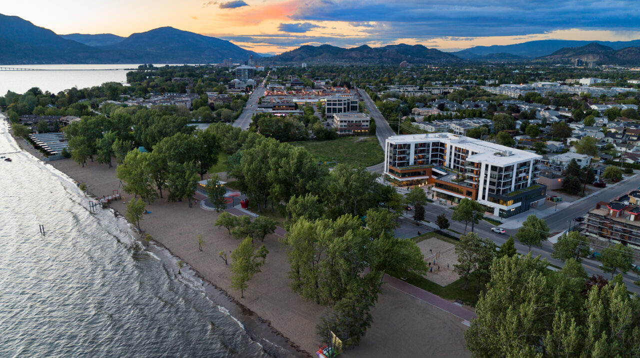 an exterior view of The Shore Kelowna, a hotel-apartment, showing Kelowna's lower mission neighbourhood, Gyro beach and Okanagan Lake in the foreground