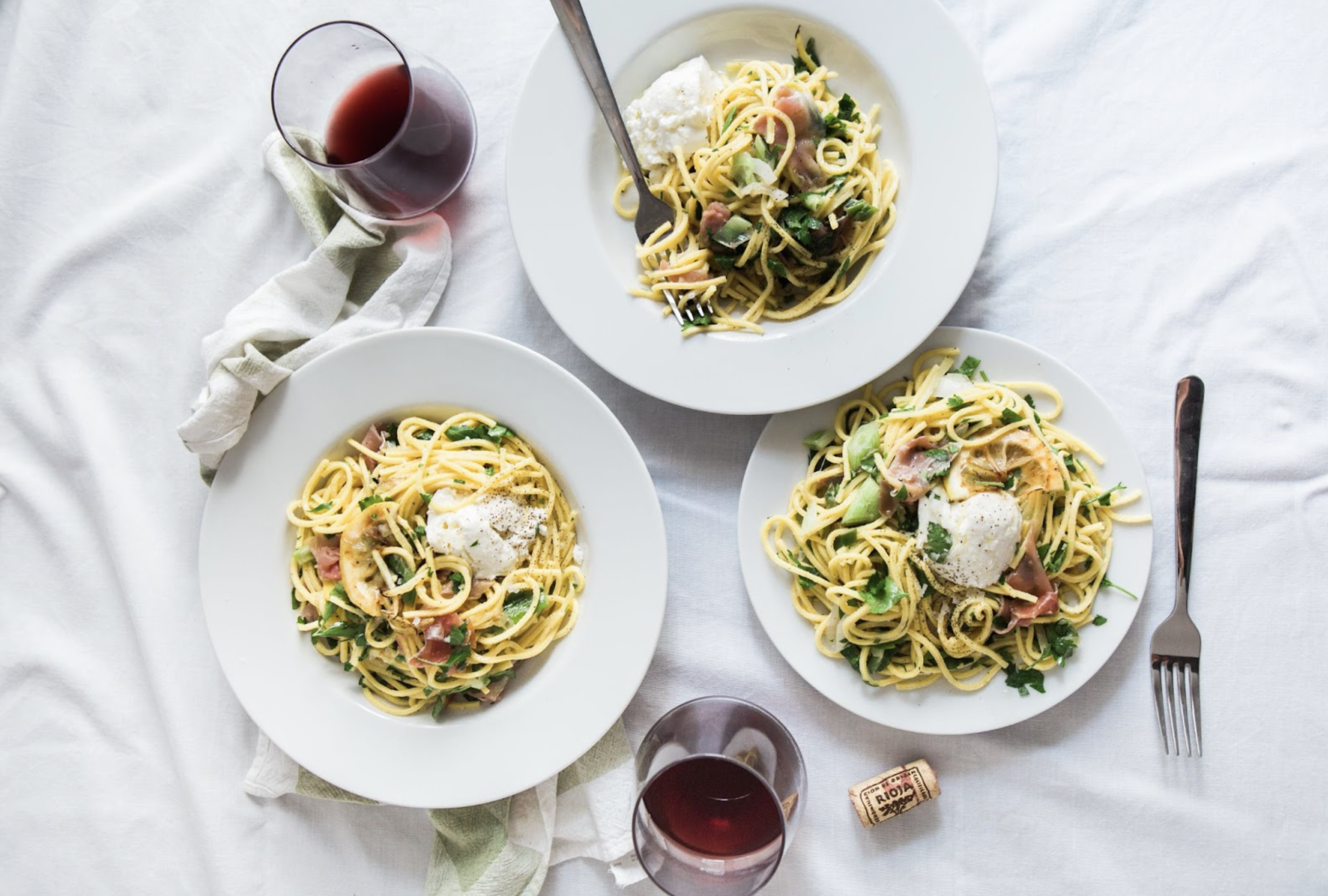 a top-down view of an italian restaurant meal of pasta in three plates, with two glasses of wine over a white tablecloth