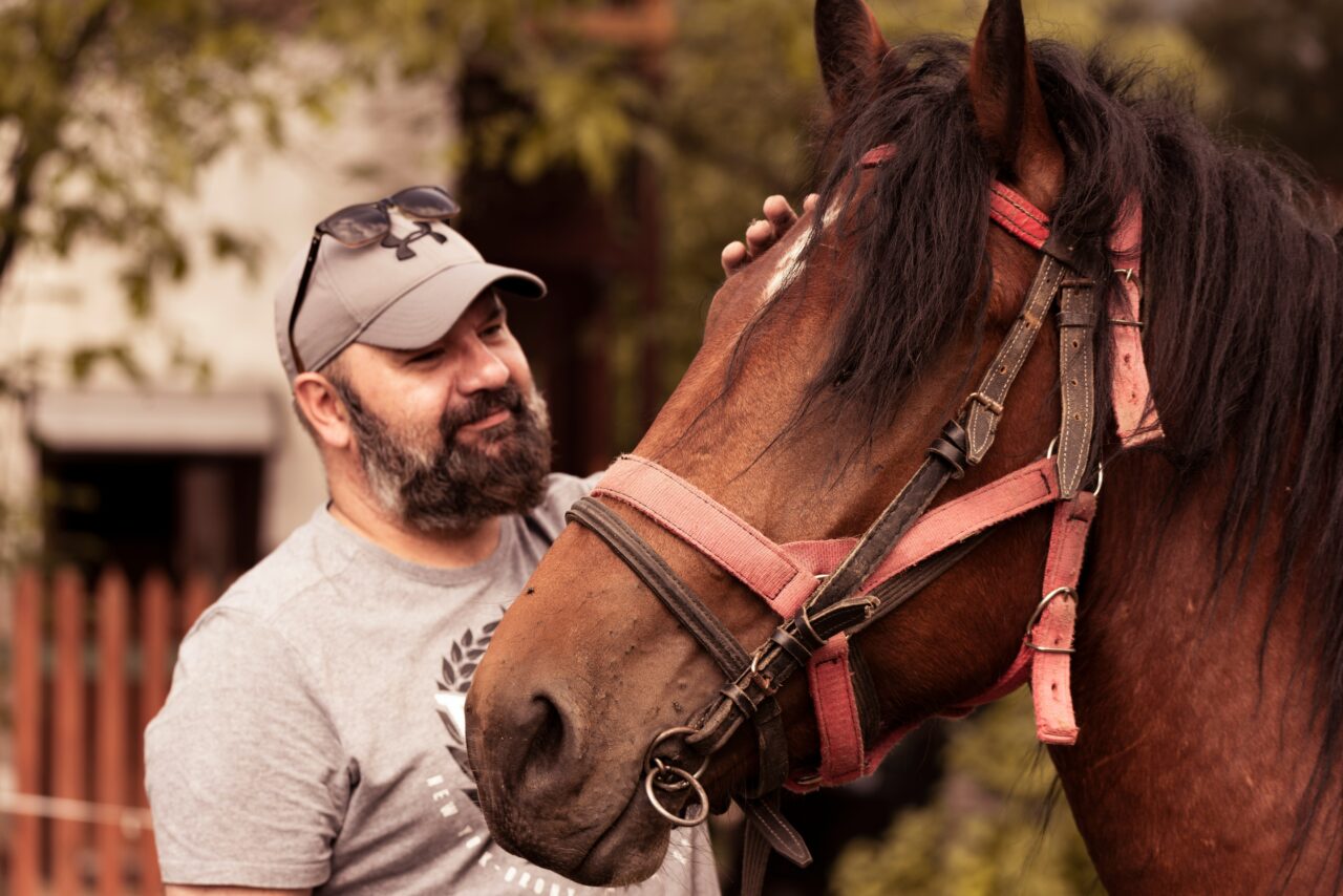 A bearded man in a grey hat and grey shirt standing with a brown horse petting its head.