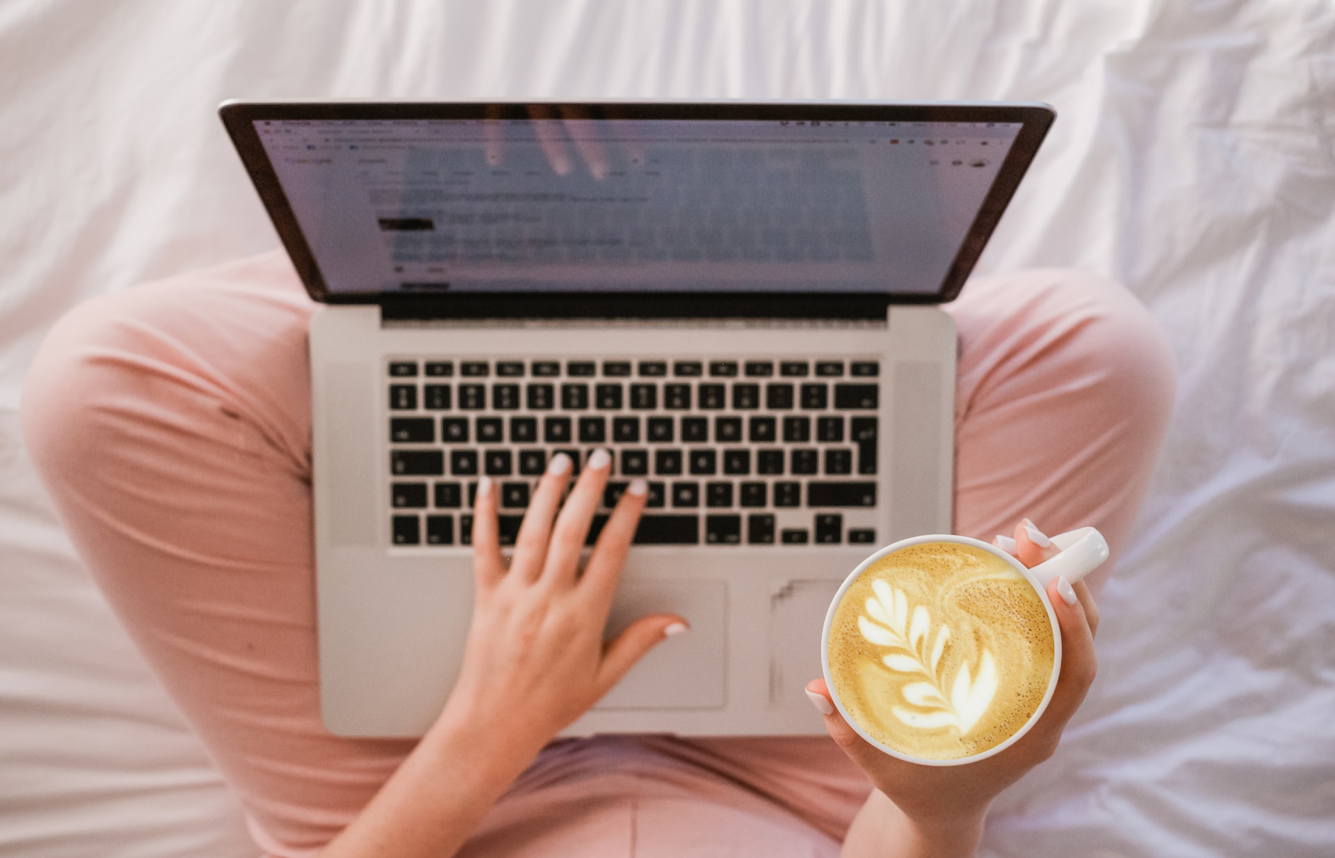 a top-down view of a woman typing on a laptop, with the laptop on her lap sitting cross legged on white sheets, holding a latte with leaf foam art