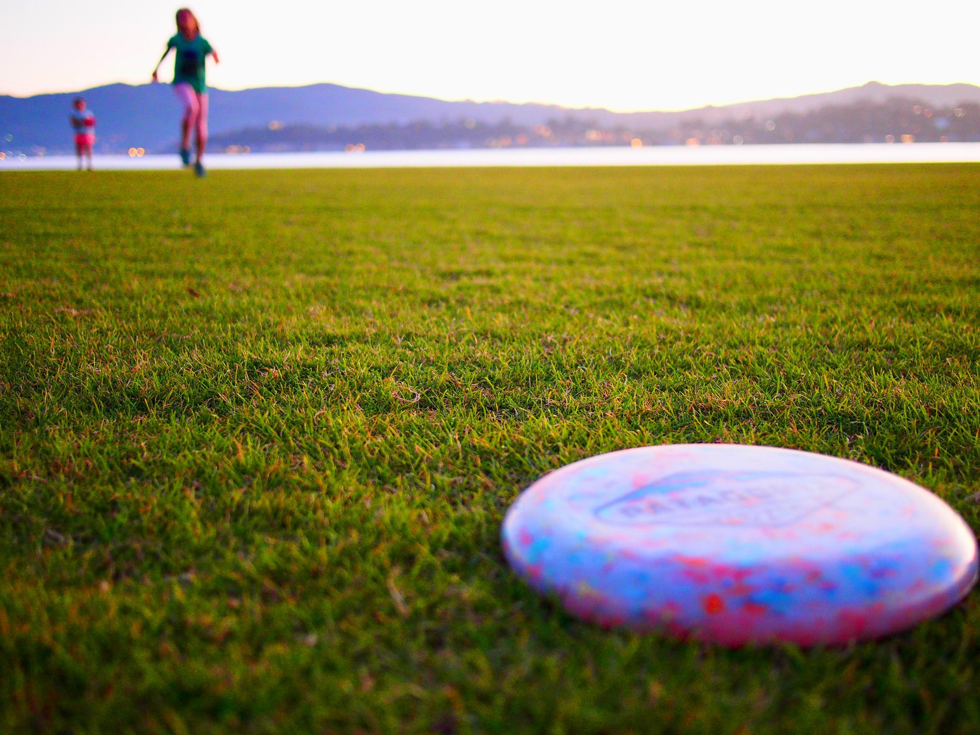 A closeup of a frisbee lying on the grass at dusk with two kids running towards it at a Kelowna park