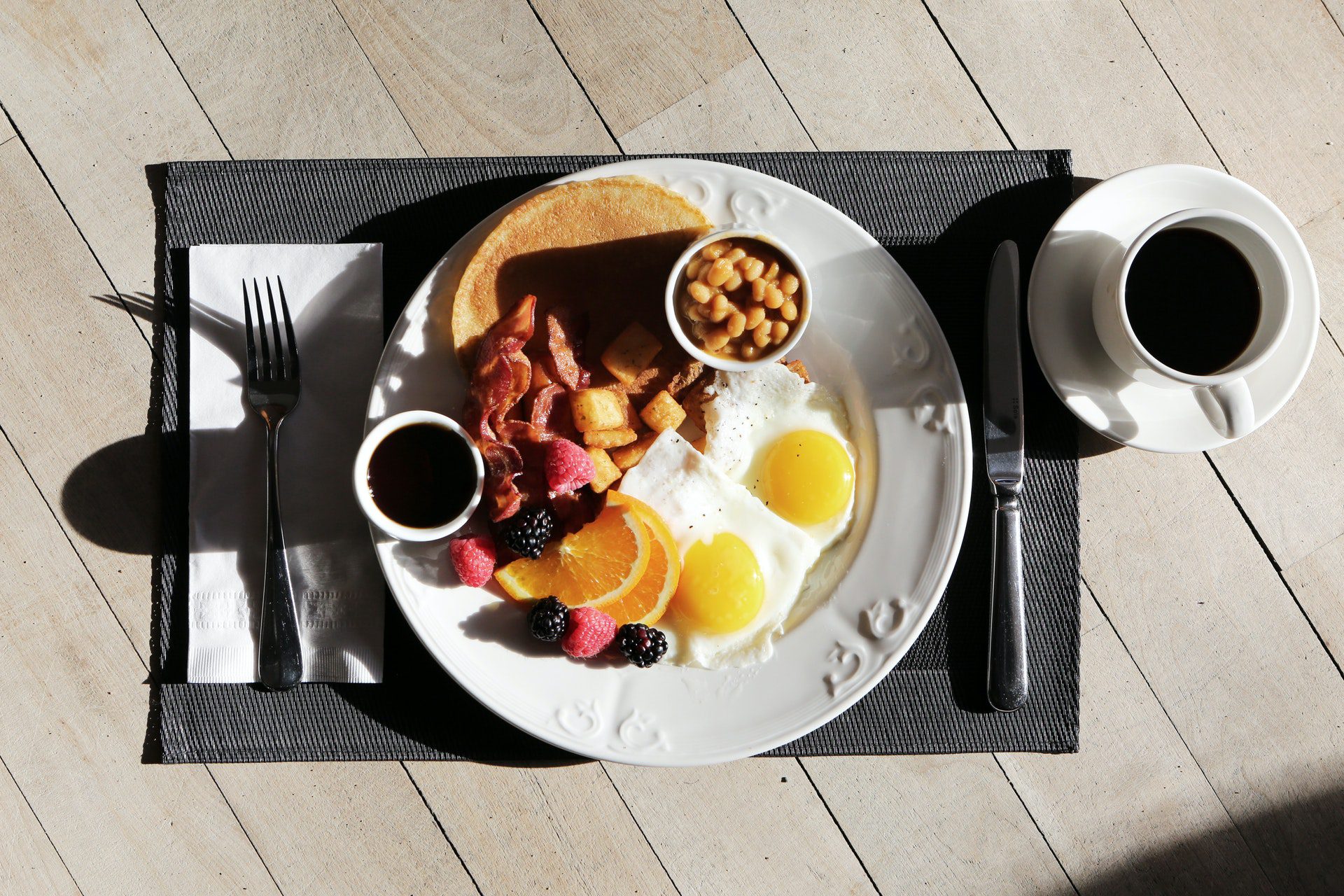 a breakfast setting on a wooden table with a plate of eggs, pancakes, bacon and fruits