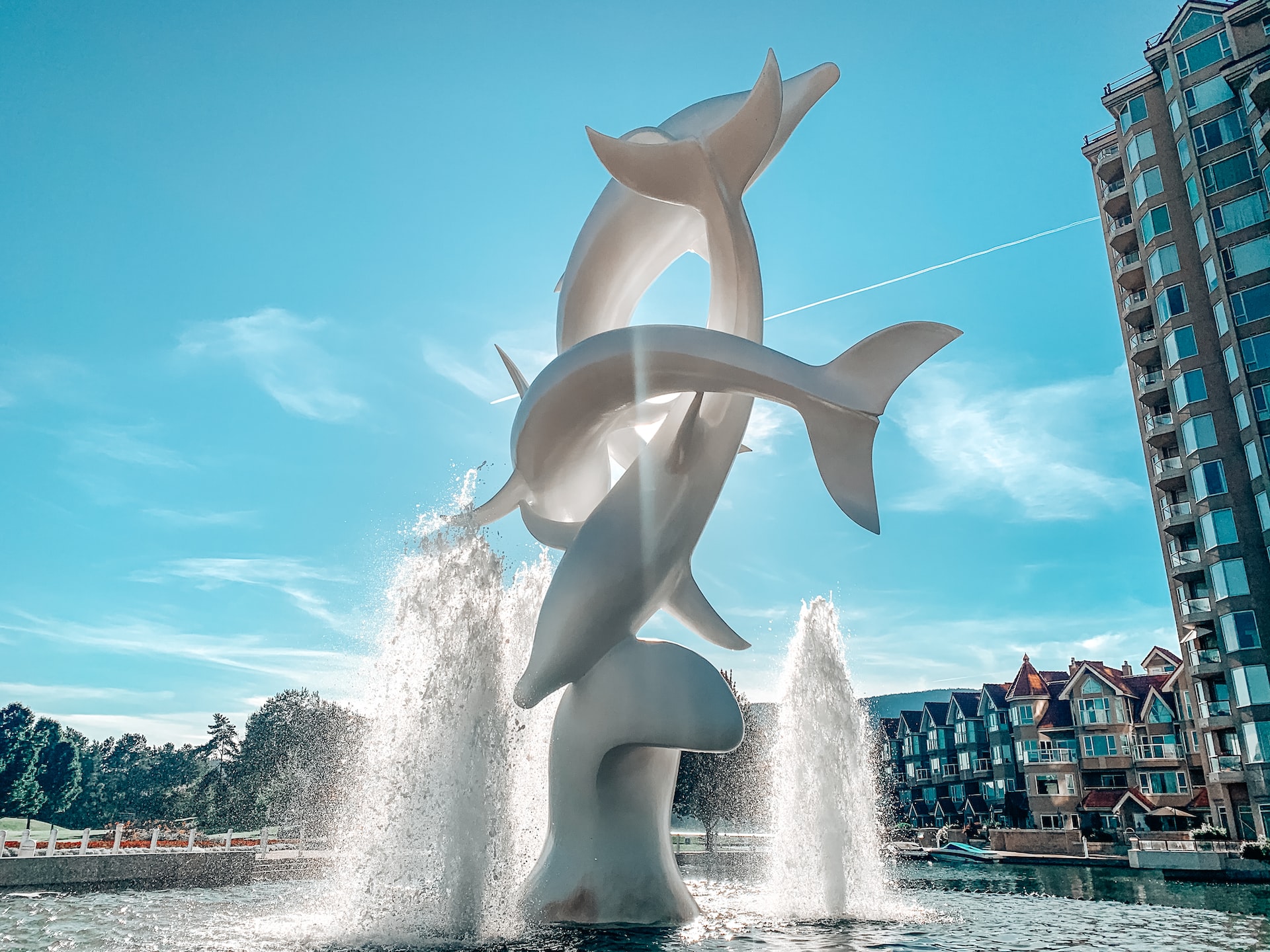 a life size sculpture called the dolphins in downtown kelowna showing three dolphins jumping in white marble with a fountain surrounding; behind is the Delta Grand hotel, included in this guide for booking kelowna hotels