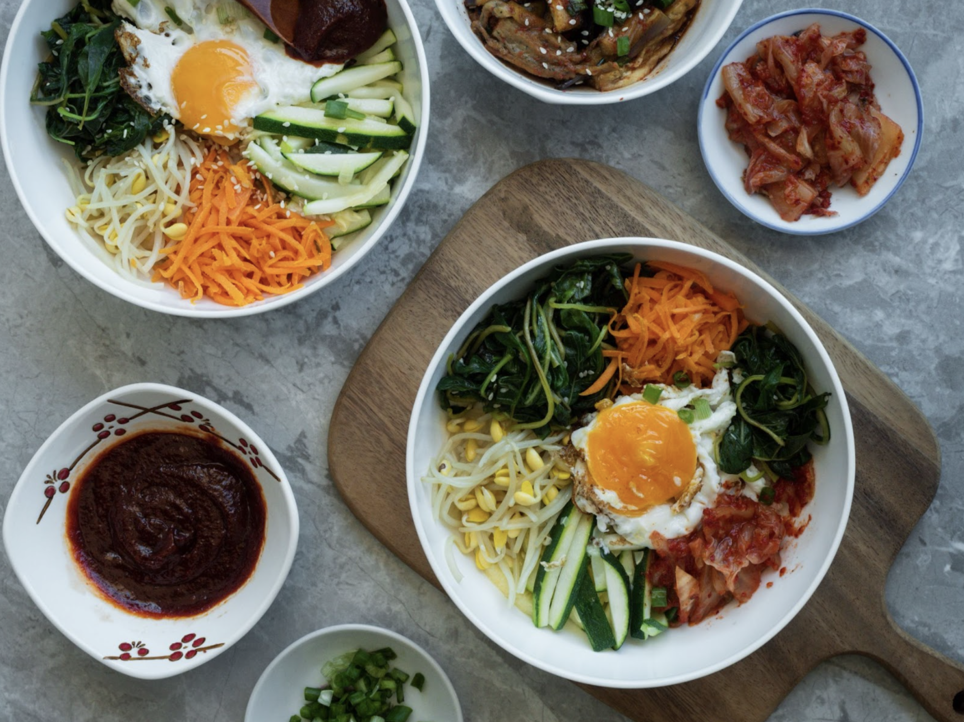 top-down view of bowls of Kelowna's best Korean food, with side sauces, on a granite countertop with a wooden cutting board under one bowl